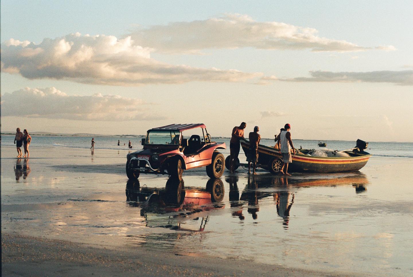 A red dune buggy and a boat are on a wet beach with several people standing near them and in the water under a cloudy sky.