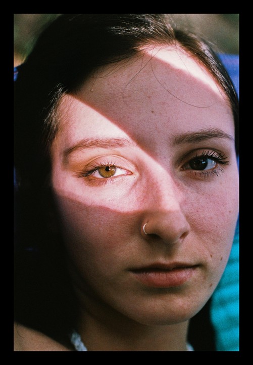 A woman with a nose ring looks directly at the viewer, with a diagonal shadow across her face.