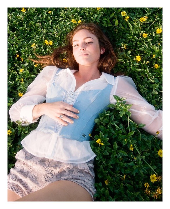 A woman with brown hair lies in a field of green plants with small yellow flowers.