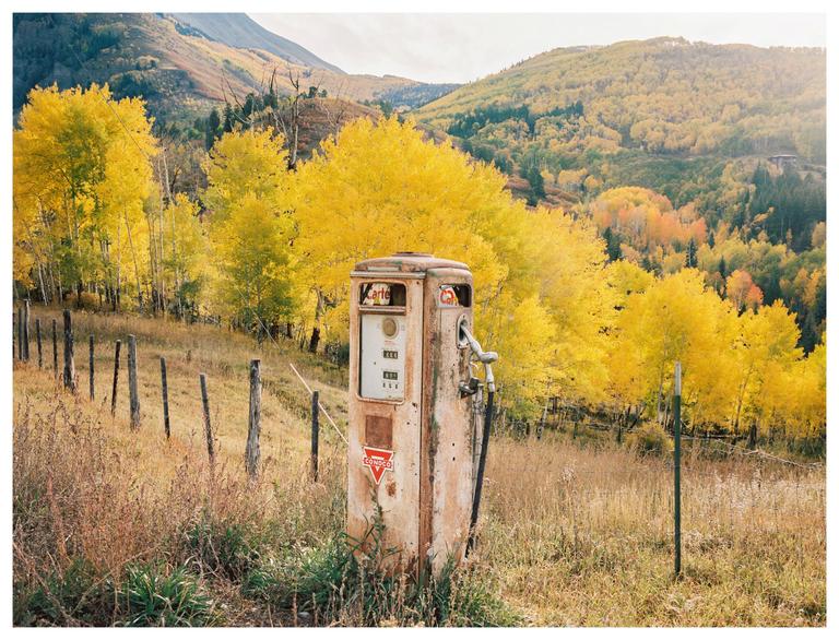 An old gas pump stands in a field with a mountain and autumn trees in the background.