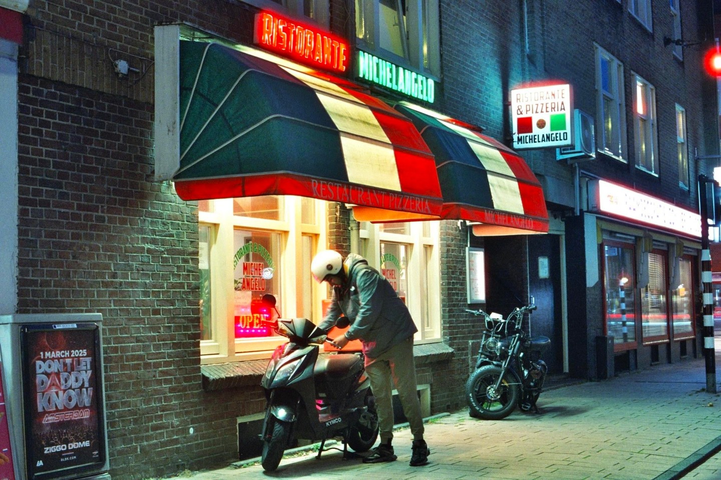 A person wearing a helmet stands next to a scooter parked in front of a restaurant with a striped awning at night.