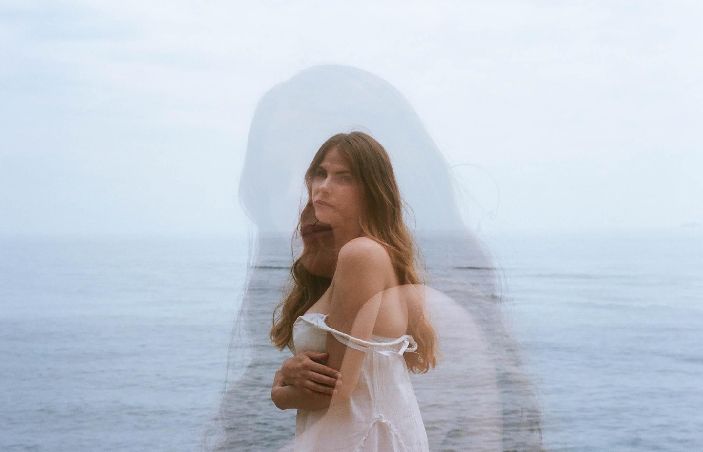 A woman with long hair stands in front of the ocean with a double exposure effect.