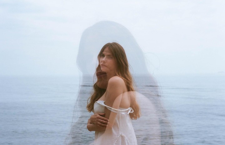 A woman with long hair stands in front of the ocean with a double exposure effect.