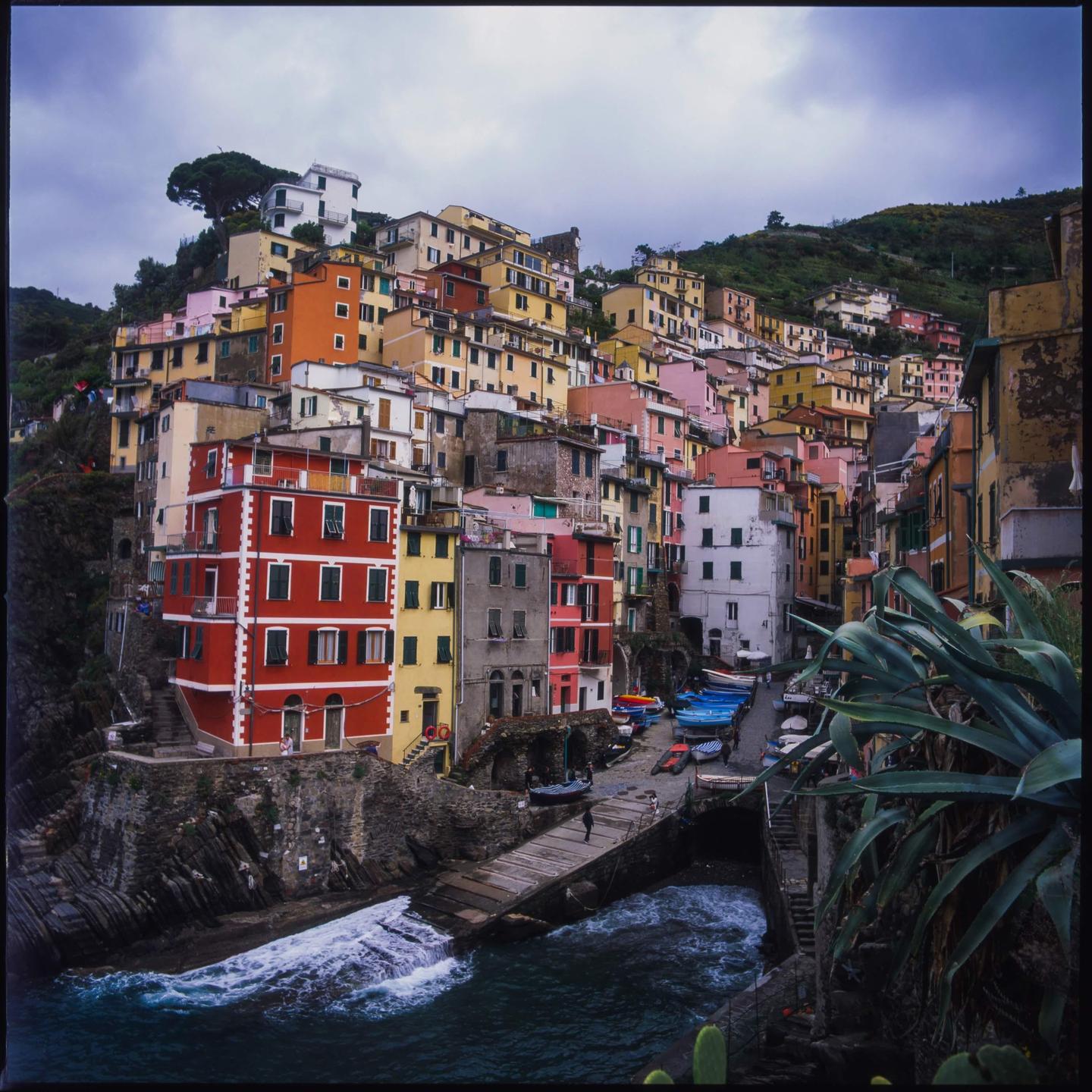 A colorful village with houses built into a hillside overlooks a small harbor with boats and crashing waves.
