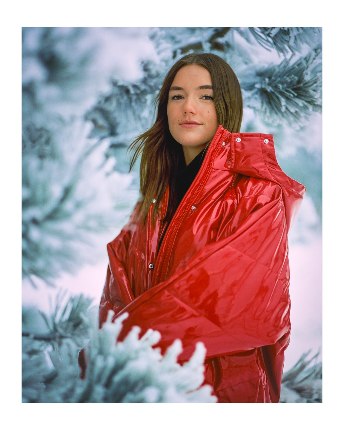 A woman in a shiny red jacket stands among snow-covered pine branches.