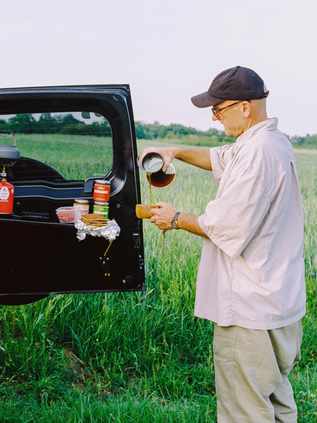 A man pours coffee from a metal pot into a wooden cup next to an open car trunk in a grassy field.