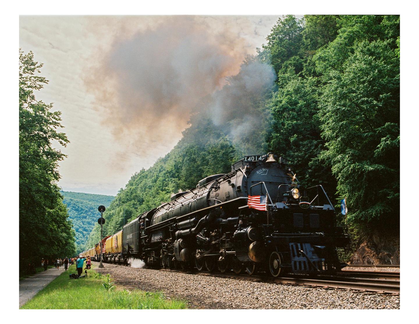 A large black steam locomotive with an American flag on its front travels along a track next to a forested hillside.
