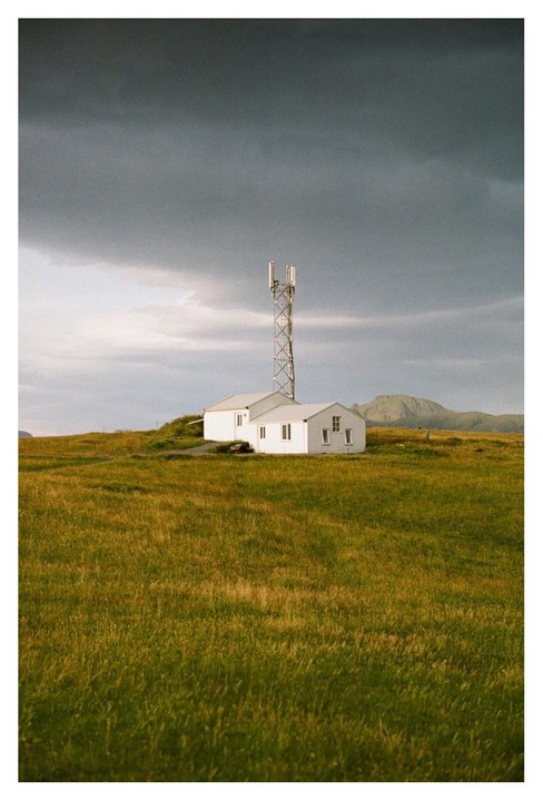 A white building with a tall antenna stands in a field under a cloudy sky.