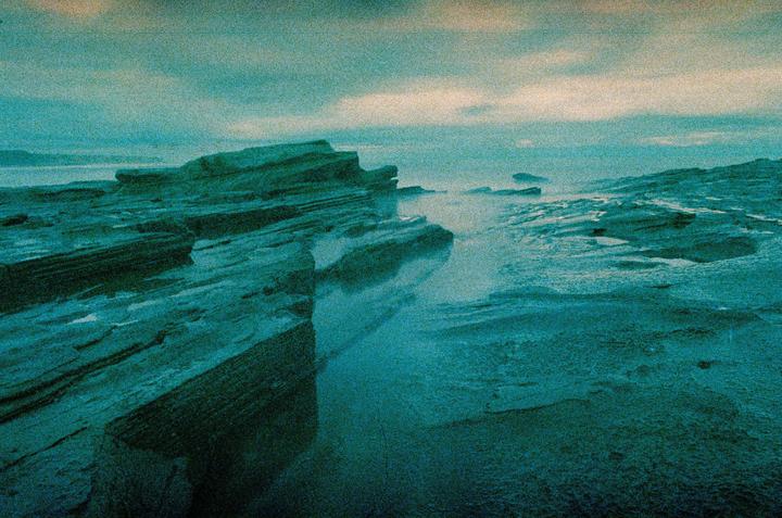 Jagged rock formations line a misty, teal-colored coastline under a cloudy sky.
