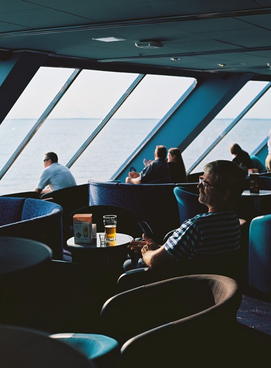 People sit in a lounge with large windows overlooking the sea.
