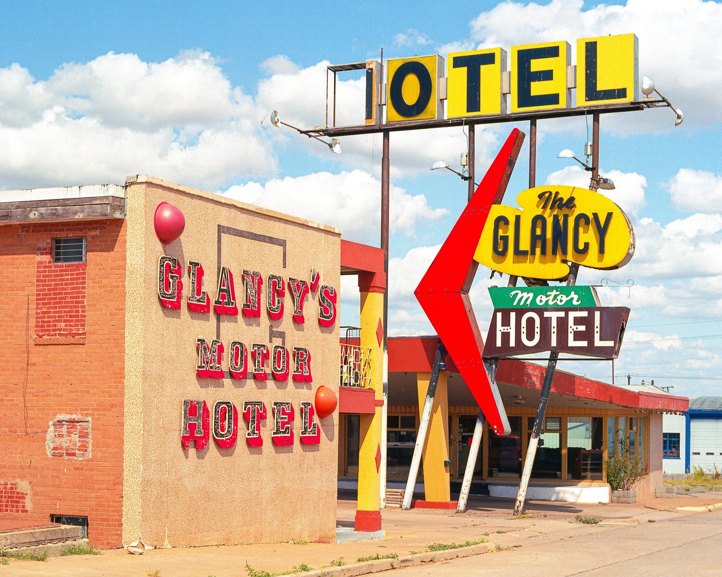 A vintage motel sign and building with "Glancy's Motor Hotel" written on them stand under a bright blue sky with white clouds.
