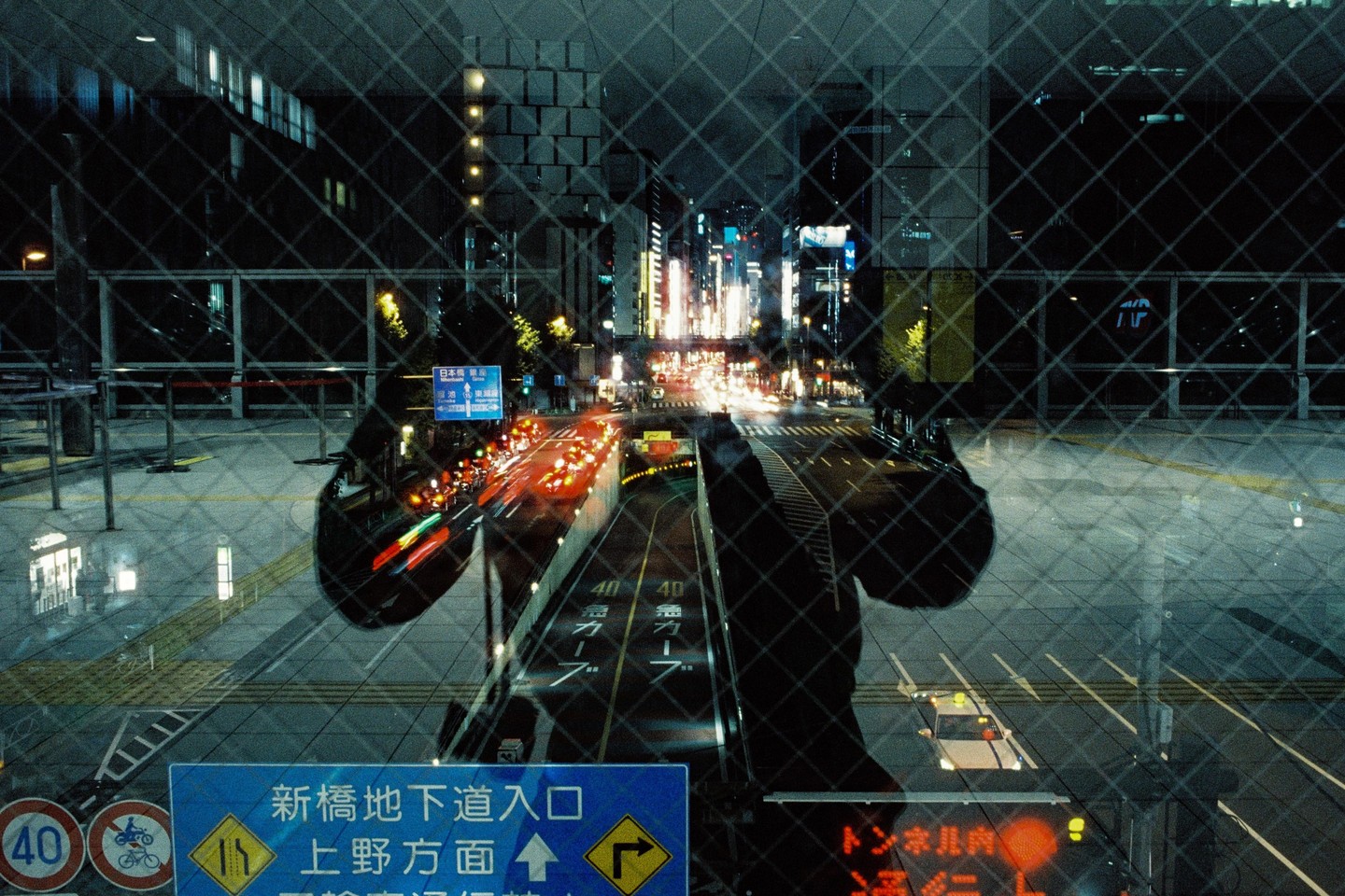 A double exposure shows a city street at night with light trails from vehicles and a reflection of a person in a window.