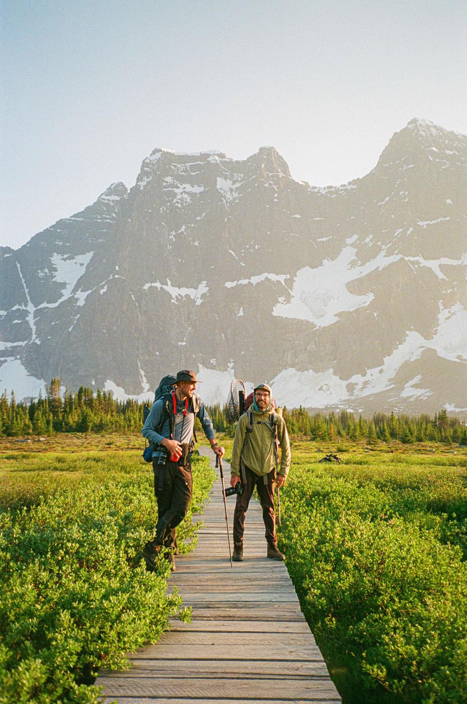Two men with backpacks and hiking poles stand on a wooden boardwalk in a field with a snow-capped mountain in the background.