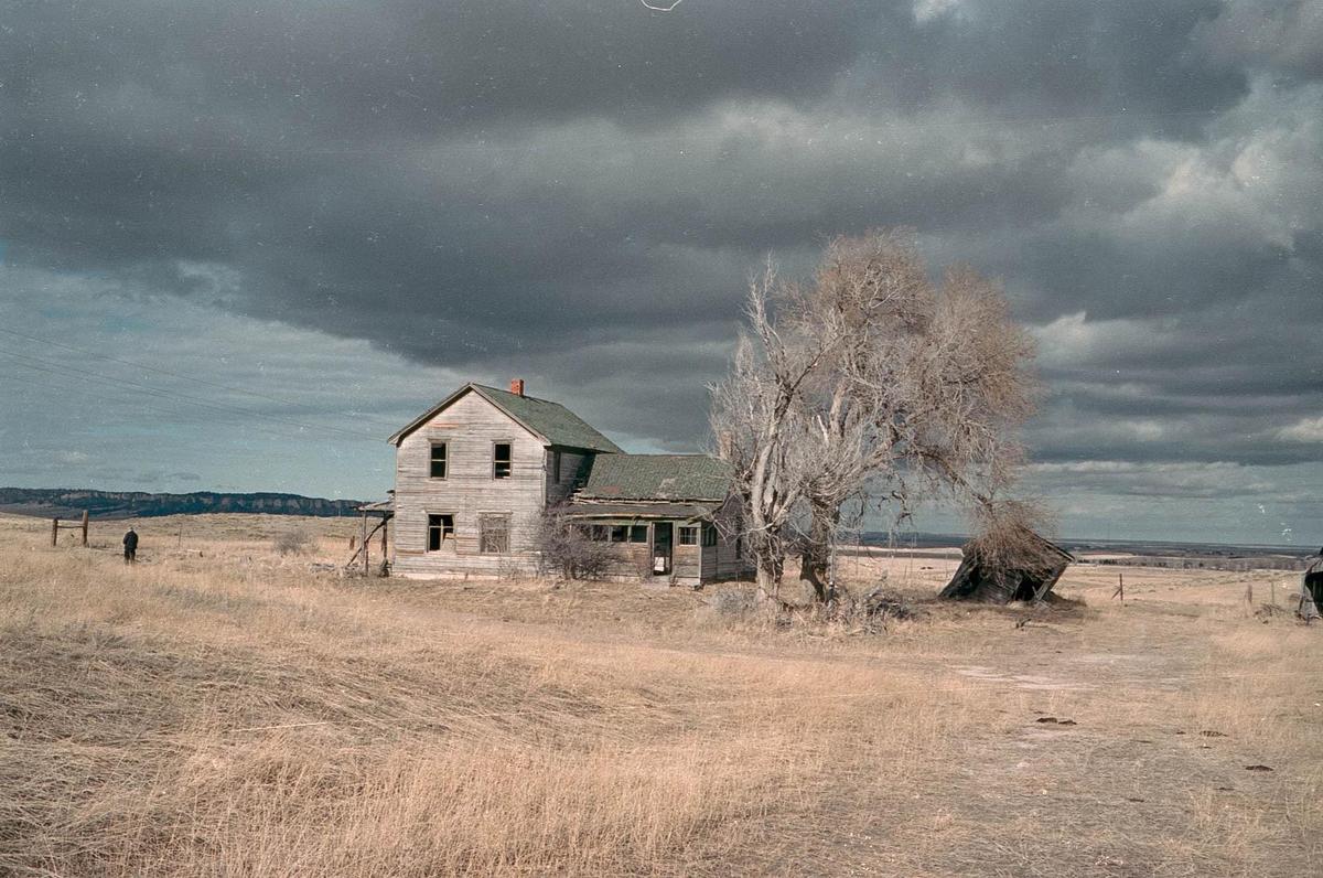 An abandoned house stands in a field under a dramatic cloudy sky, with a lone figure in the distance.