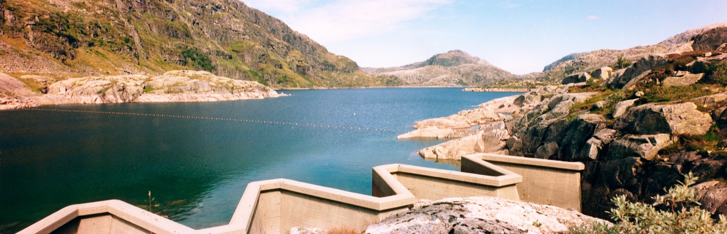 A panoramic landscape shows a large lake surrounded by rocky mountains and a concrete structure in the foreground.