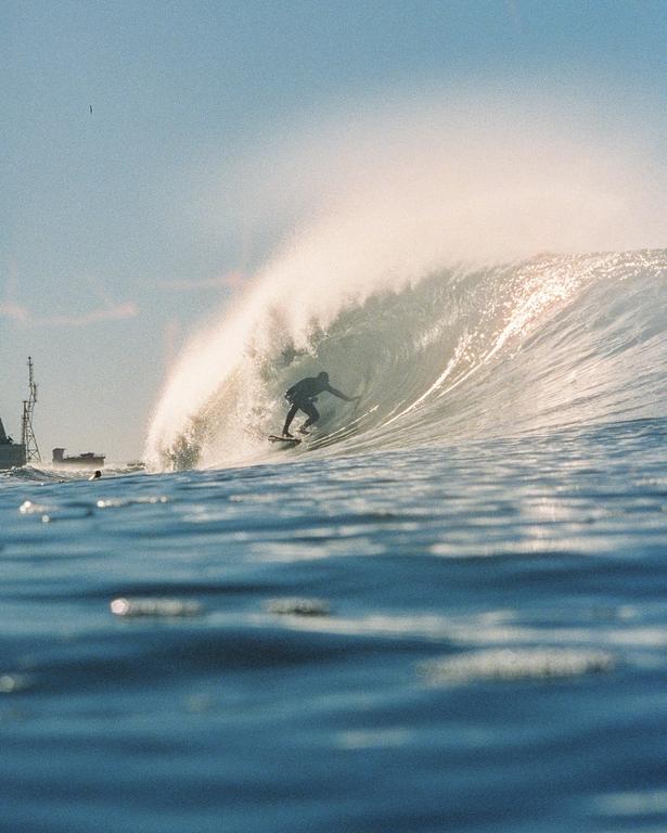 A surfer rides a large wave with a ship visible in the background.