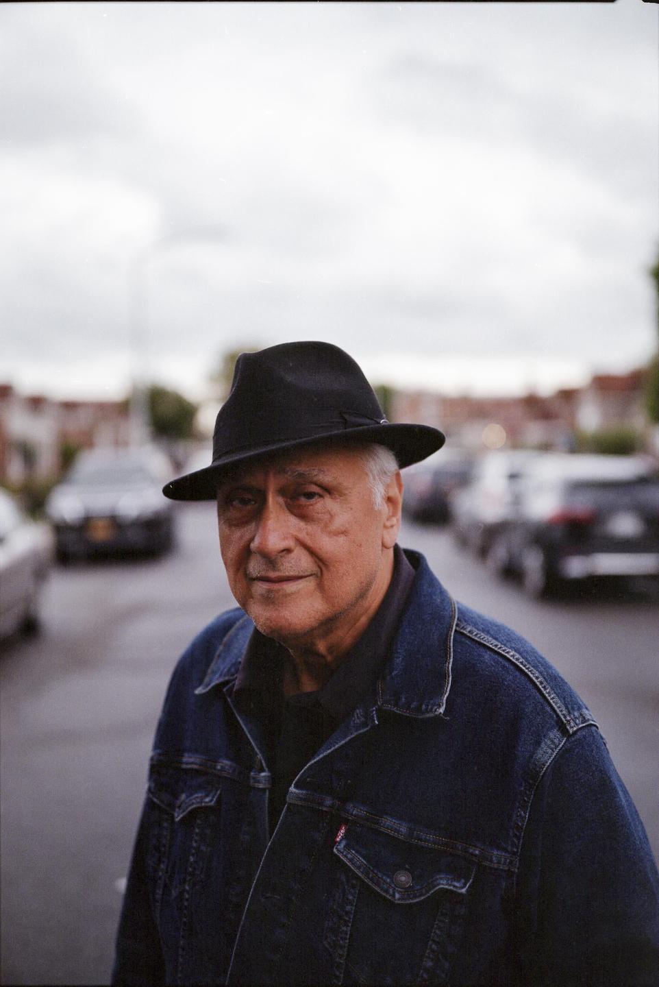A man wearing a hat and a denim jacket stands on a street with cars parked on both sides.