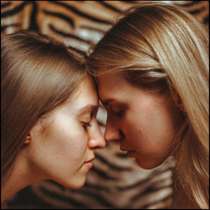 Two women with closed eyes touch foreheads against a blurred background.