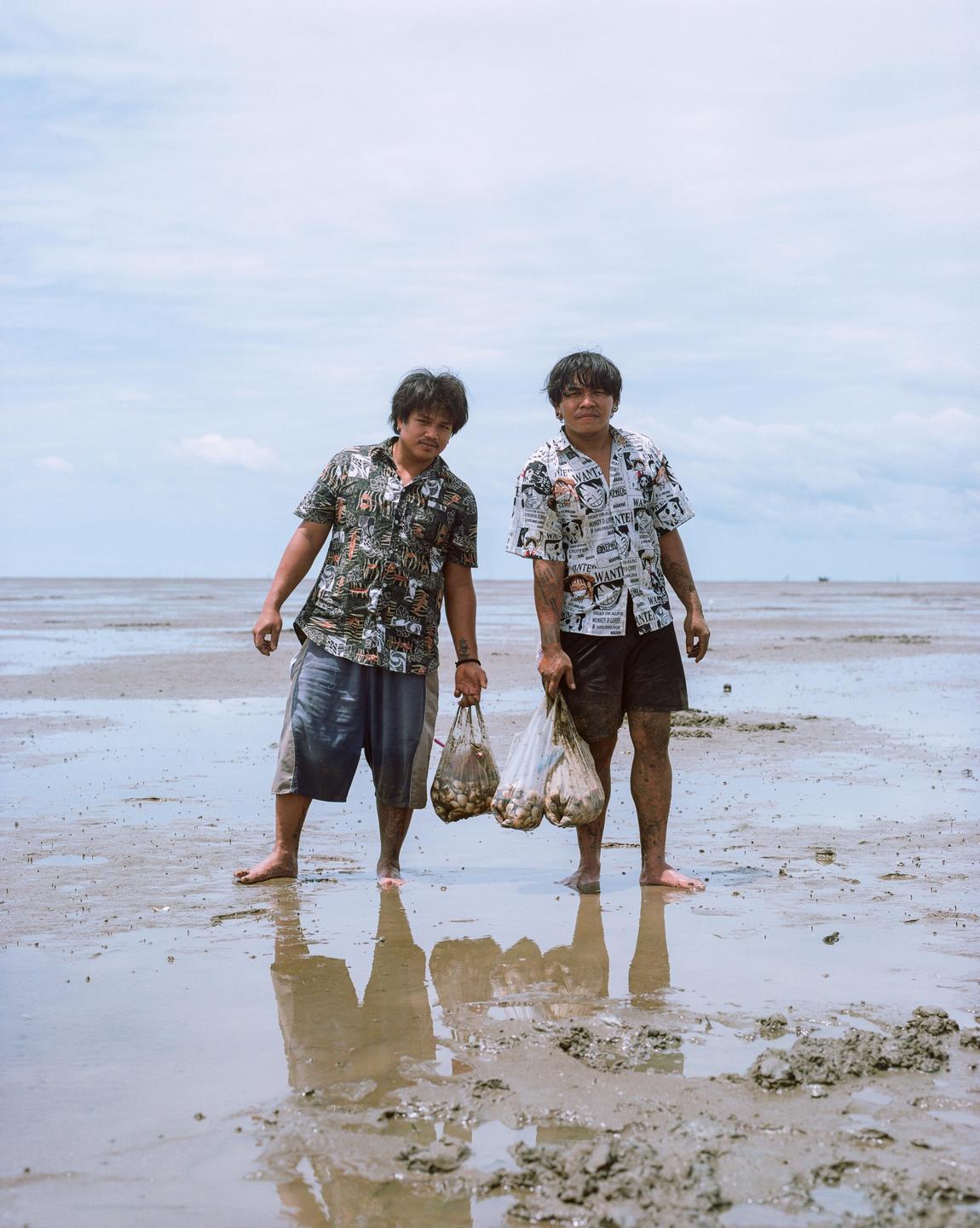 Two men stand barefoot in the mud holding bags of shellfish on a cloudy day.