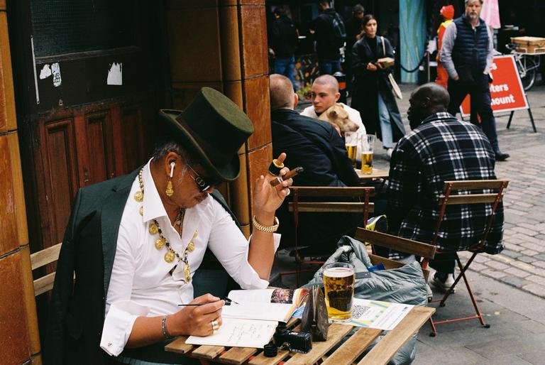 A man wearing a top hat and gold jewelry sits at an outdoor table, writing in a notebook while holding a cigar.