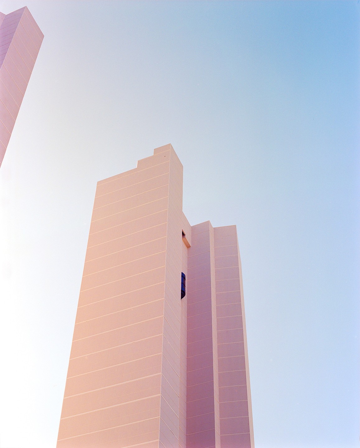 A tall, pink building with horizontal lines is viewed from below against a clear blue sky.
