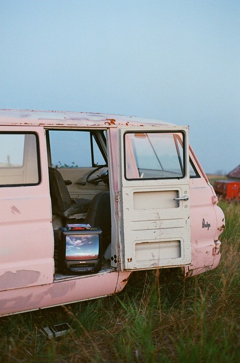 A pink and white van with open doors sits in a grassy field with a television set inside showing a cloudy sky.