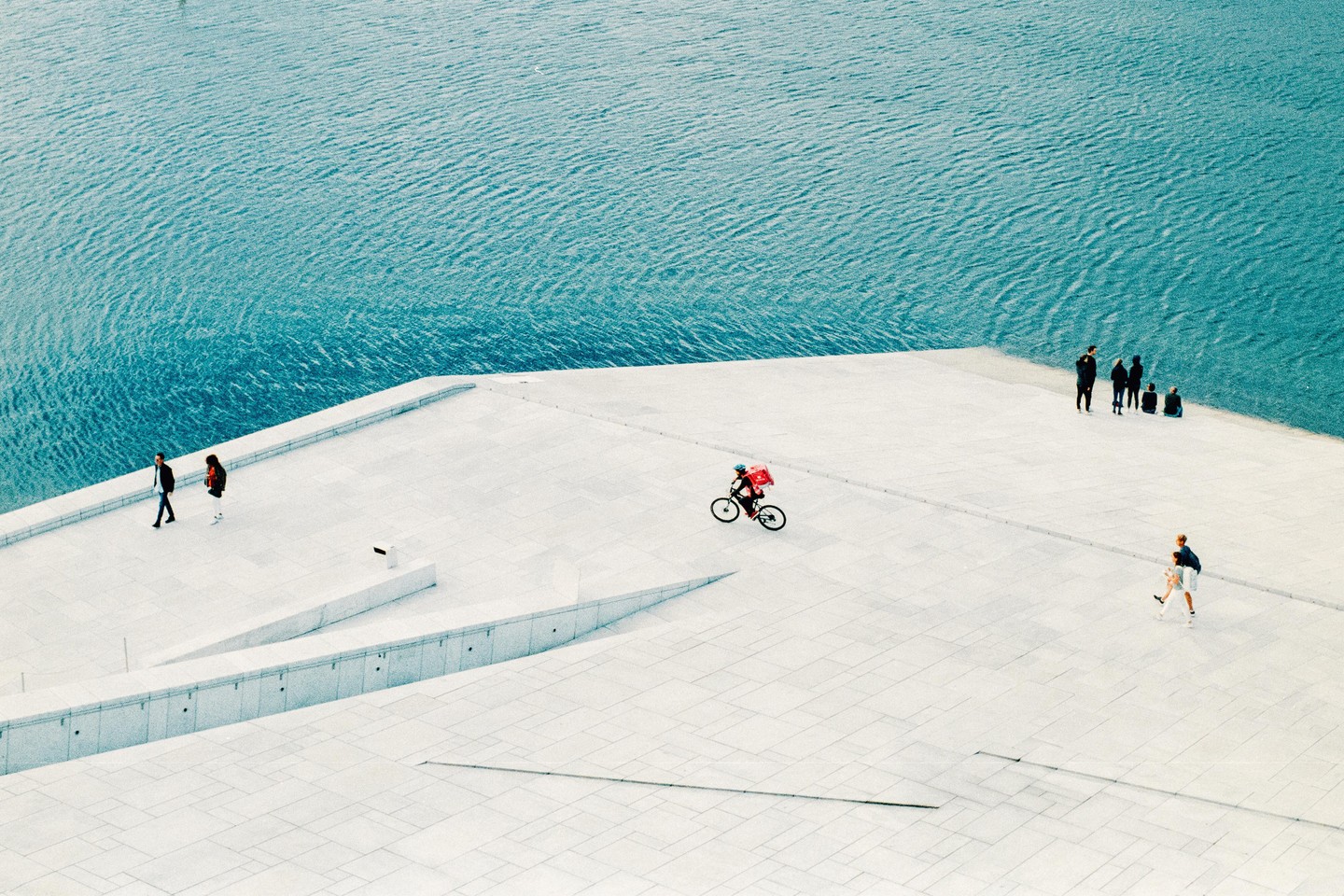 A high-angle view shows people walking and cycling on the white, angular architecture of the Oslo Opera House next to blue water.