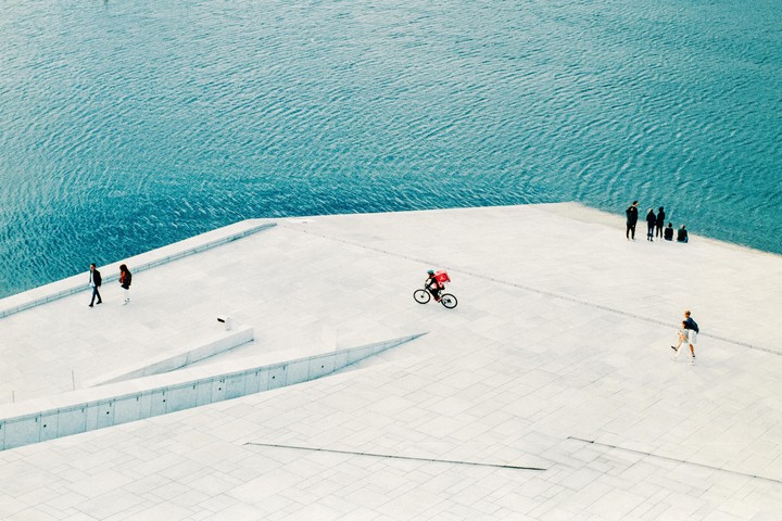 A high-angle view shows people walking and cycling on the white, angular architecture of the Oslo Opera House next to blue water.