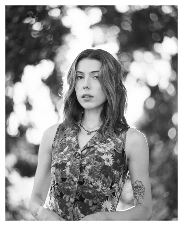 A woman with wavy hair and a floral top stands outdoors, looking directly at the viewer.