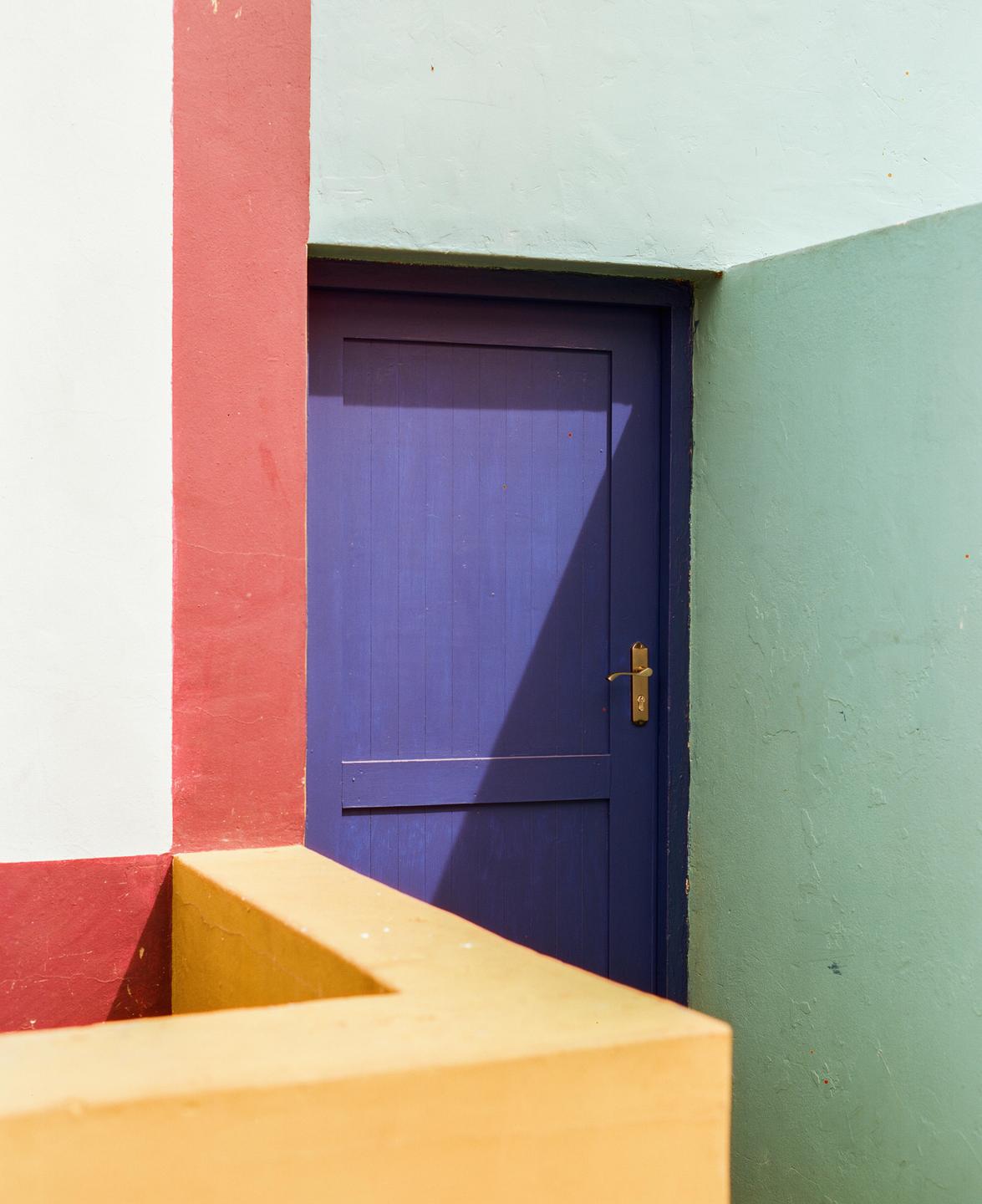 A purple door is set into a wall with red, white, and mint green sections, with a yellow railing in the foreground.