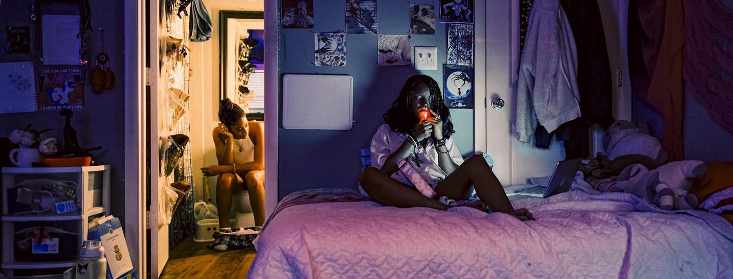 A woman sits on a bed with a laptop while another woman sits on a toilet in a bathroom.