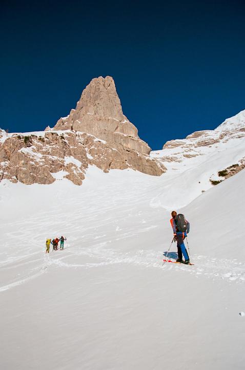 A group of skiers ascends a snowy mountain slope with a prominent rocky peak in the background under a clear blue sky.