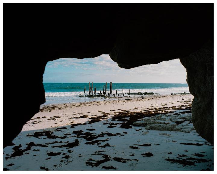 A sandy beach with seaweed and old wooden posts in the water is framed by dark rocks.