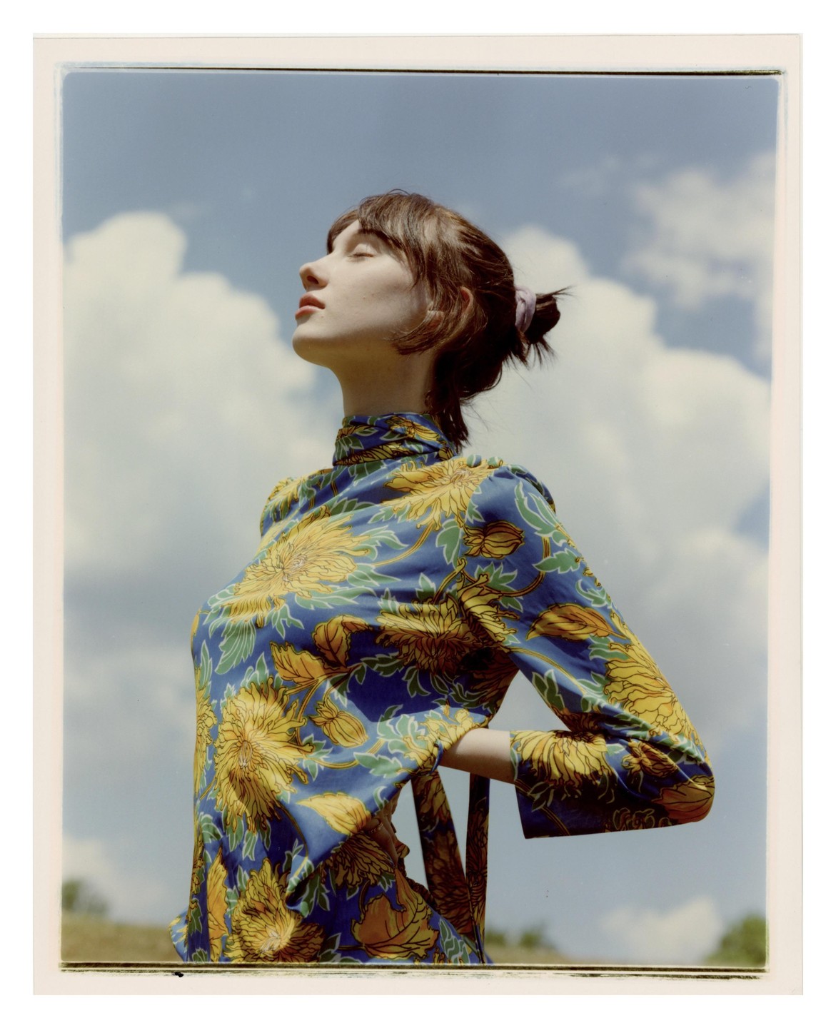 A woman with her eyes closed and face tilted towards the sky is wearing a blue and yellow floral top.