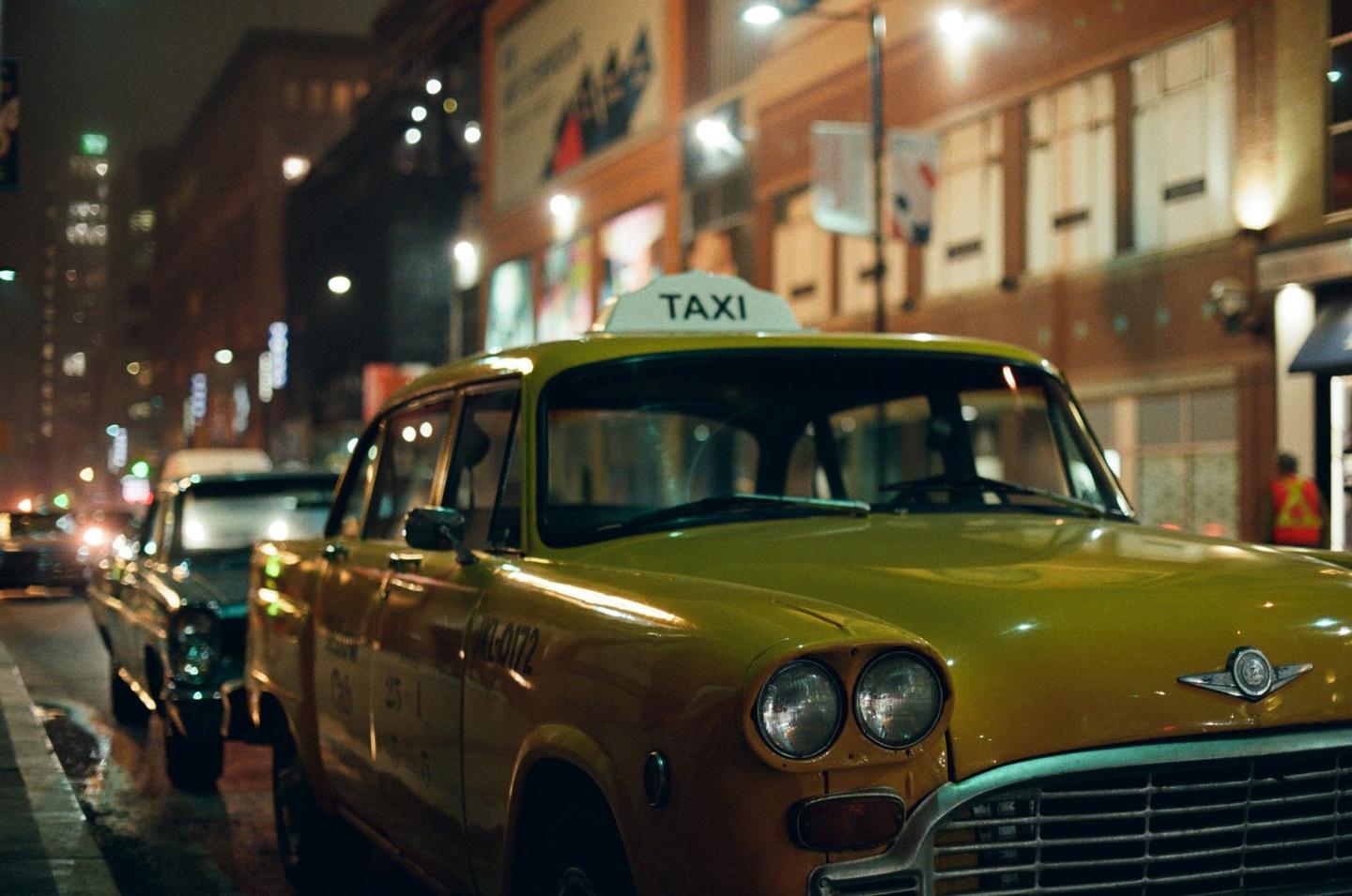 A yellow taxi is parked on a wet street at night, with city lights blurring in the background.