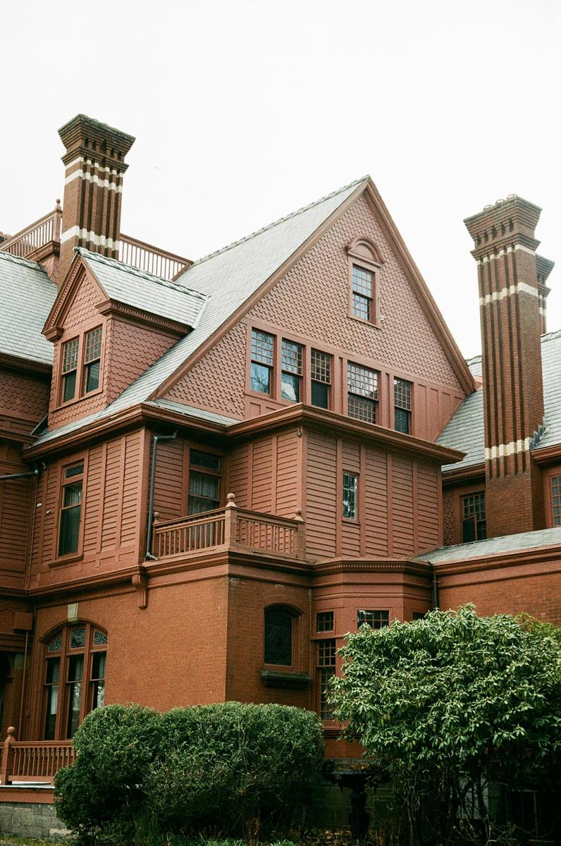 A large, ornate brick house with multiple gables and chimneys is partially obscured by green bushes.