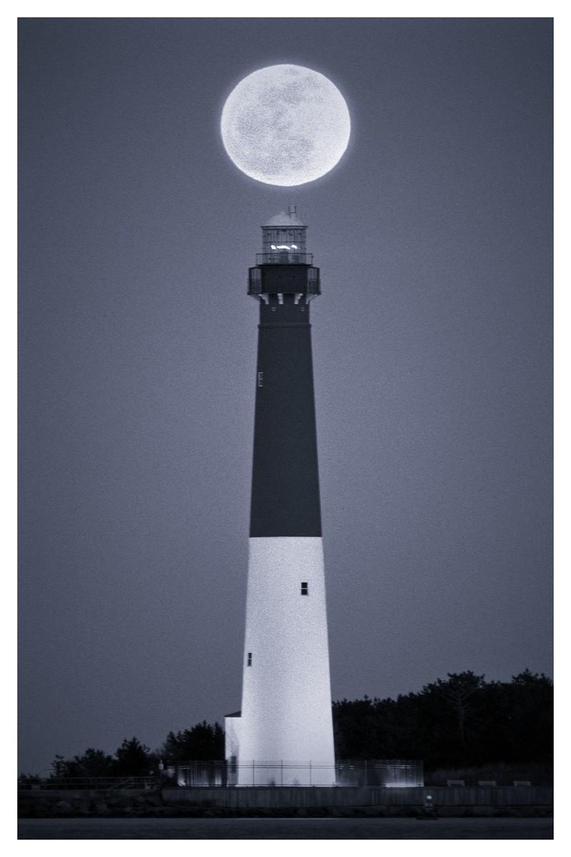 A full moon is positioned directly above a tall lighthouse at night.