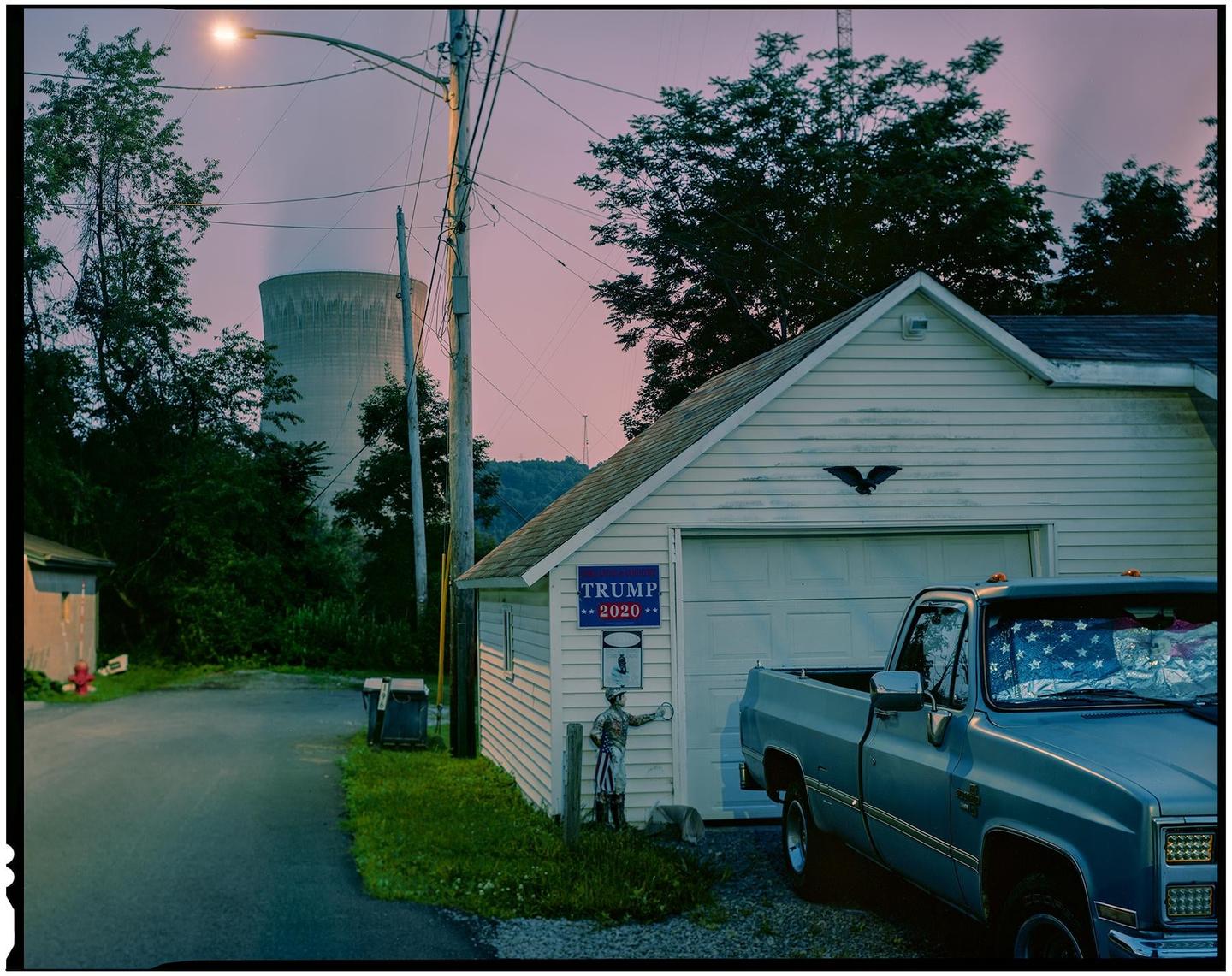 A rural street scene features a garage with a truck parked in front of it, and a power plant in the background.