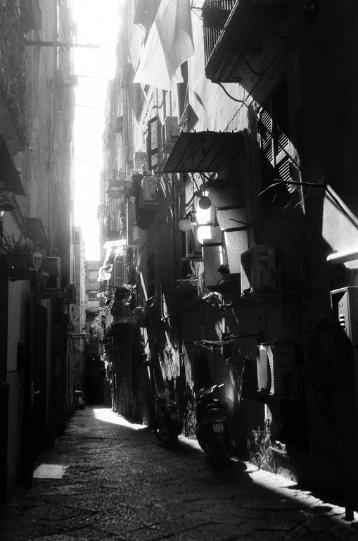 A narrow alleyway in Naples is illuminated by bright sunlight at the far end, casting deep shadows along the buildings and cobblestone street.