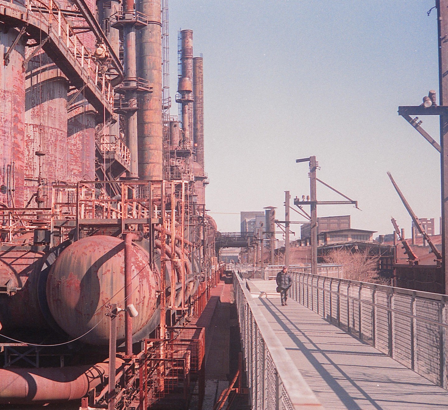A man walks along a raised walkway next to large industrial structures with multiple smokestacks.