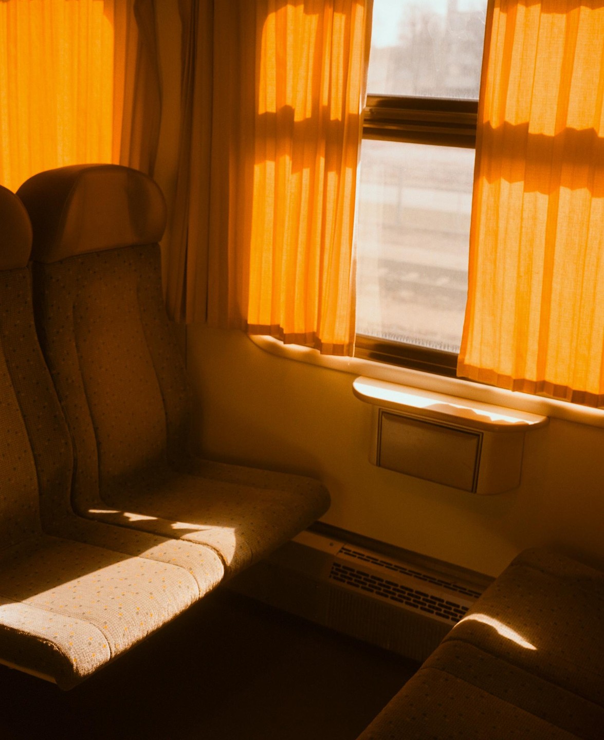 Sunlight streams through the window of a train car, illuminating two empty seats and orange curtains.