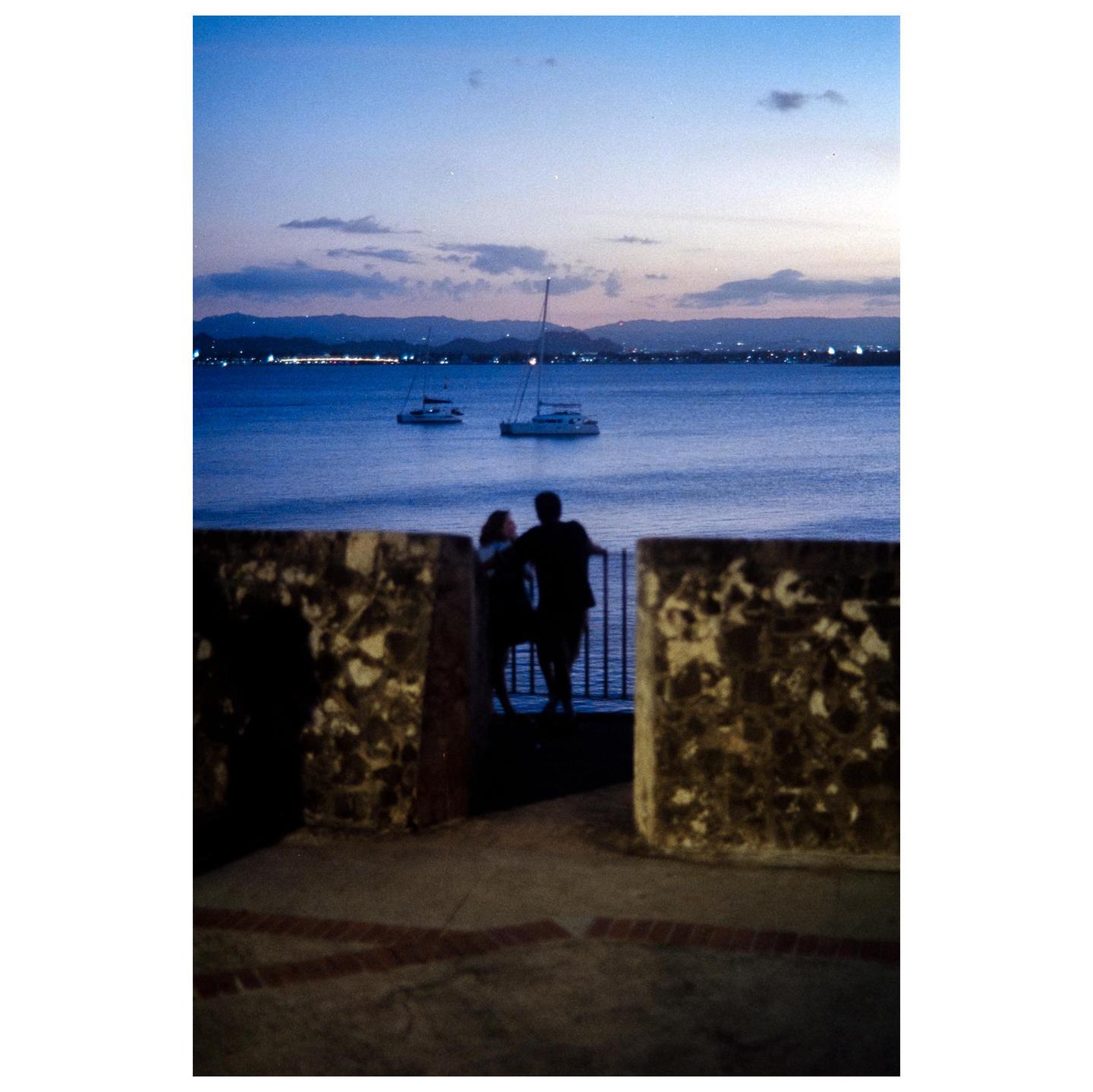 A couple stands on a stone wall overlooking a body of water with boats at sunset.