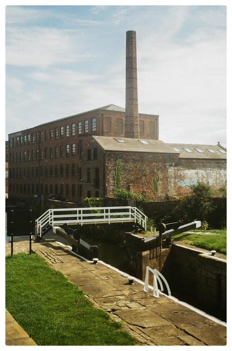 A tall brick chimney rises above a large brick building next to a canal lock with a white bridge.