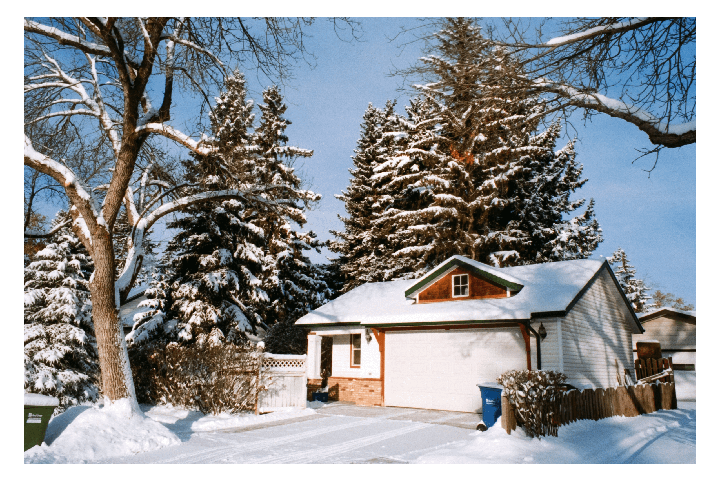 A house with a garage and a snow-covered roof is surrounded by snow-covered trees and a clear blue sky.