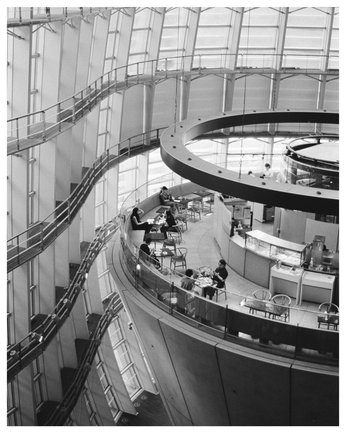 People sit at tables in a cafe with a curved balcony overlooking a large, modern interior space with a winding ramp.