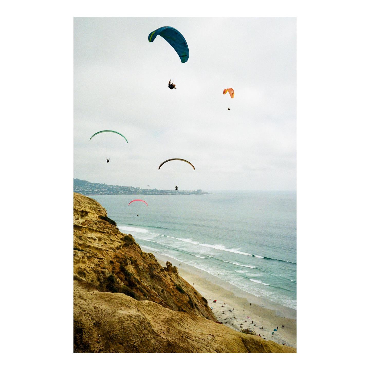 Several paragliders soar above a cliff overlooking a beach and ocean.