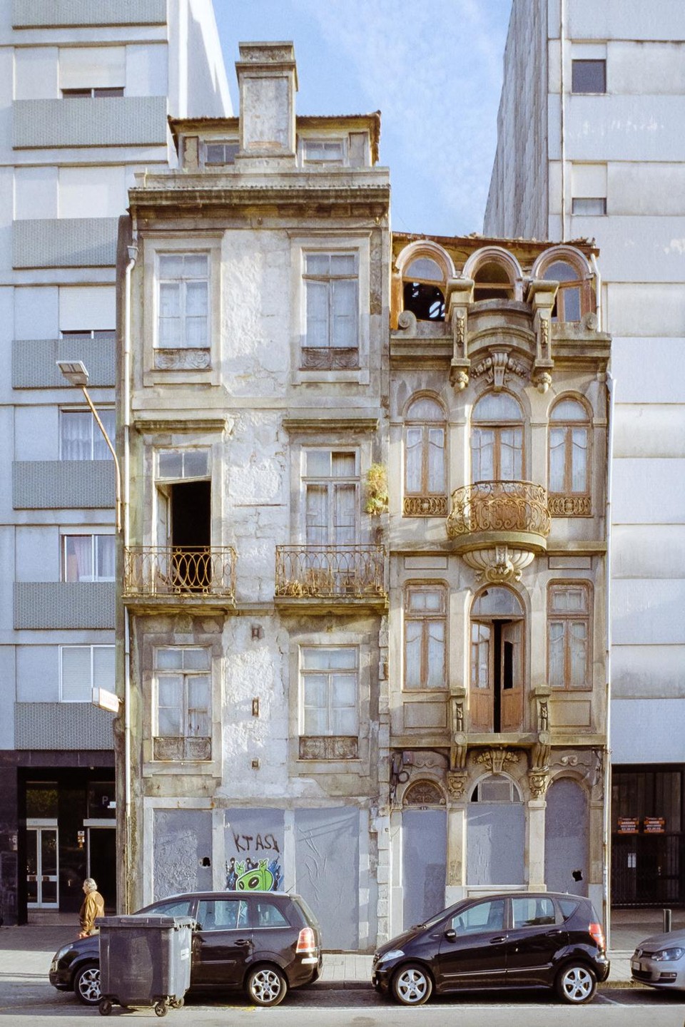 Two ornate, dilapidated buildings stand between modern, plain buildings, with cars parked on the street below.