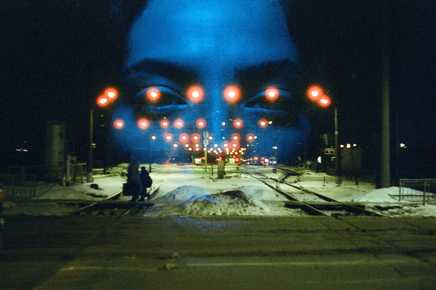 A large blue face with red lights for eyes is double exposed over a snowy street with railroad tracks at night.