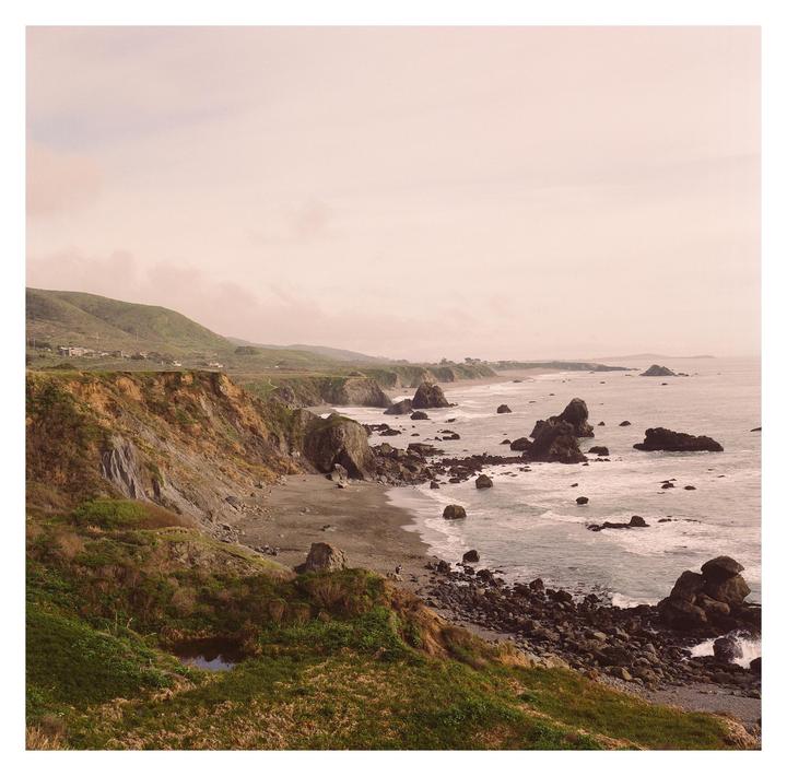 A rocky coastline with cliffs and green hills stretches along the ocean under a hazy sky.