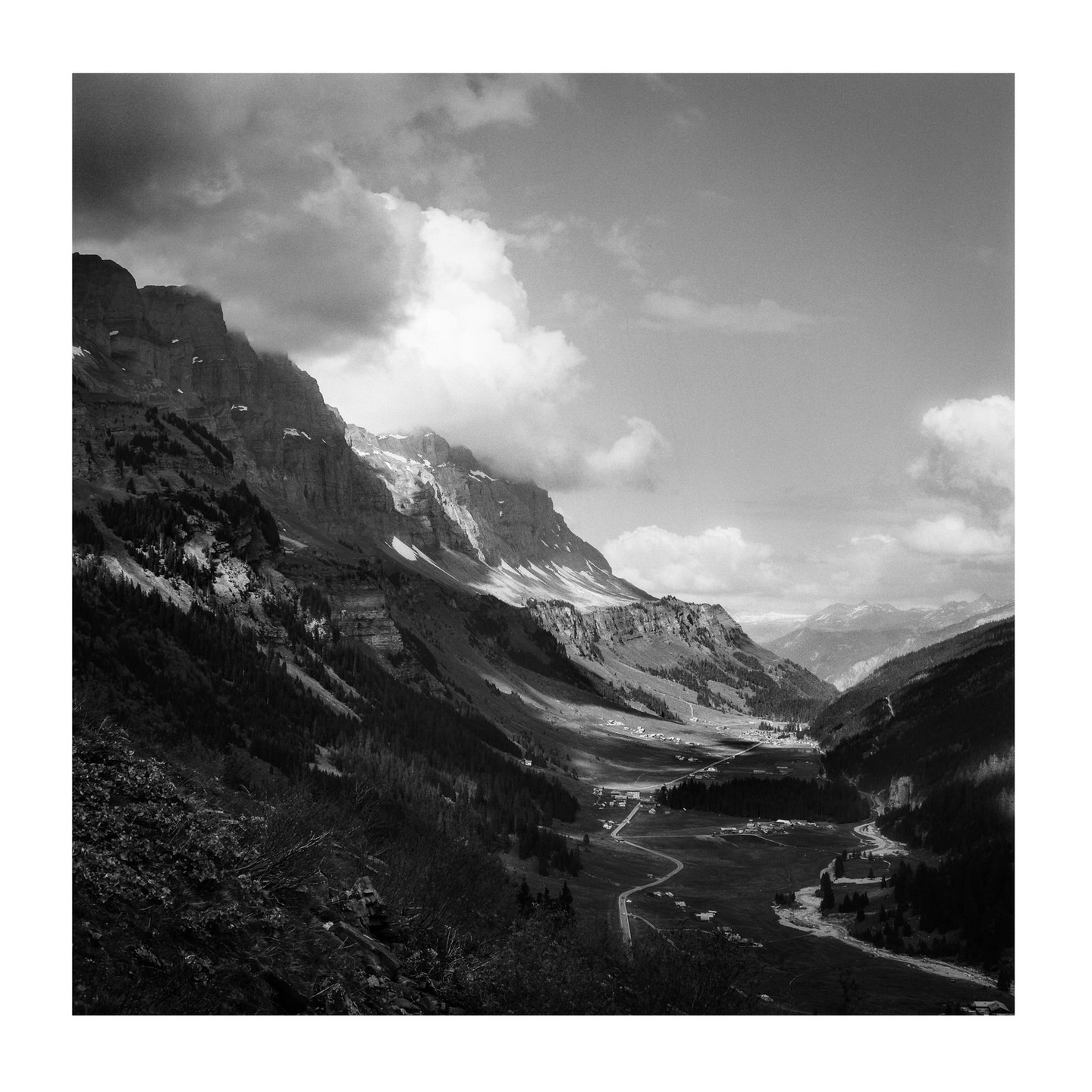 A winding road and river lead through a valley surrounded by mountains with patches of snow and clouds.